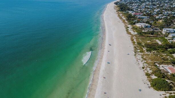 An aerial view of a beach with clear turquoise waters, a sandy shore, and a residential area with houses and greenery. - Cute and Cozy Island Bungalow, newly remodeled and a short walk to the beach (Holmes Beach)