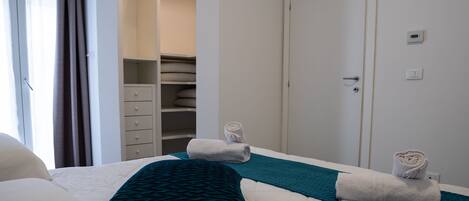 Ein Hotelzimmer mit einem Bett, einem türkisfarbenen Kopfkissen, einer weißen Decke und einem aufgerollten Handtuch auf dem Bett. Ein Schrank mit gefalteten Handtüchern und eine Tür mit Thermostat und Lichtschalter sind ebenfalls vorhanden.