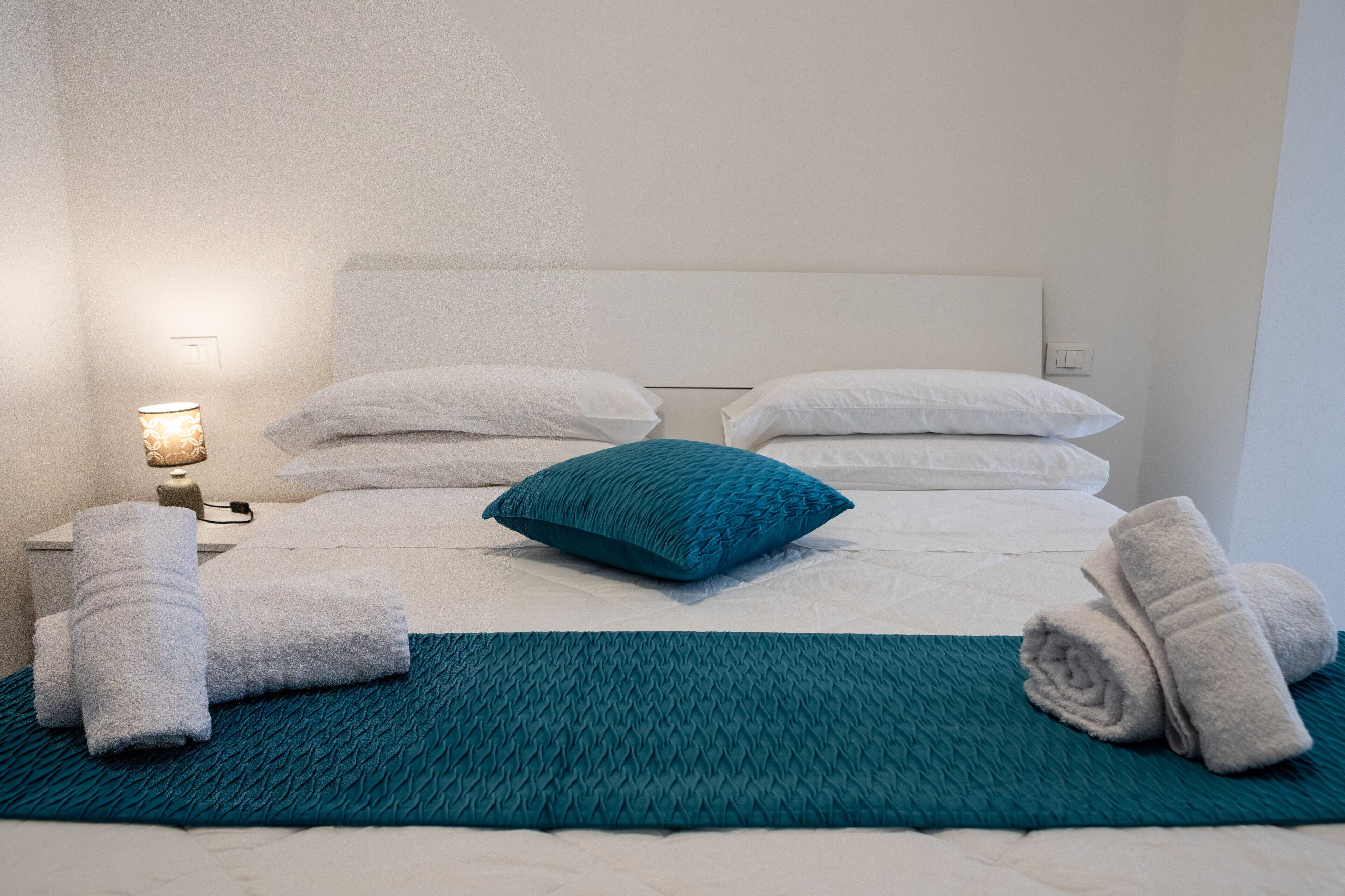 Ein ordentlich bezogenes Bett mit weißen Leinentüchern, einer türkisfarbenen Tagesdecke und einem türkisfarbenen Kissen. Auf jeder Seite des Bettes liegen zwei zusammengerollte Handtücher.
