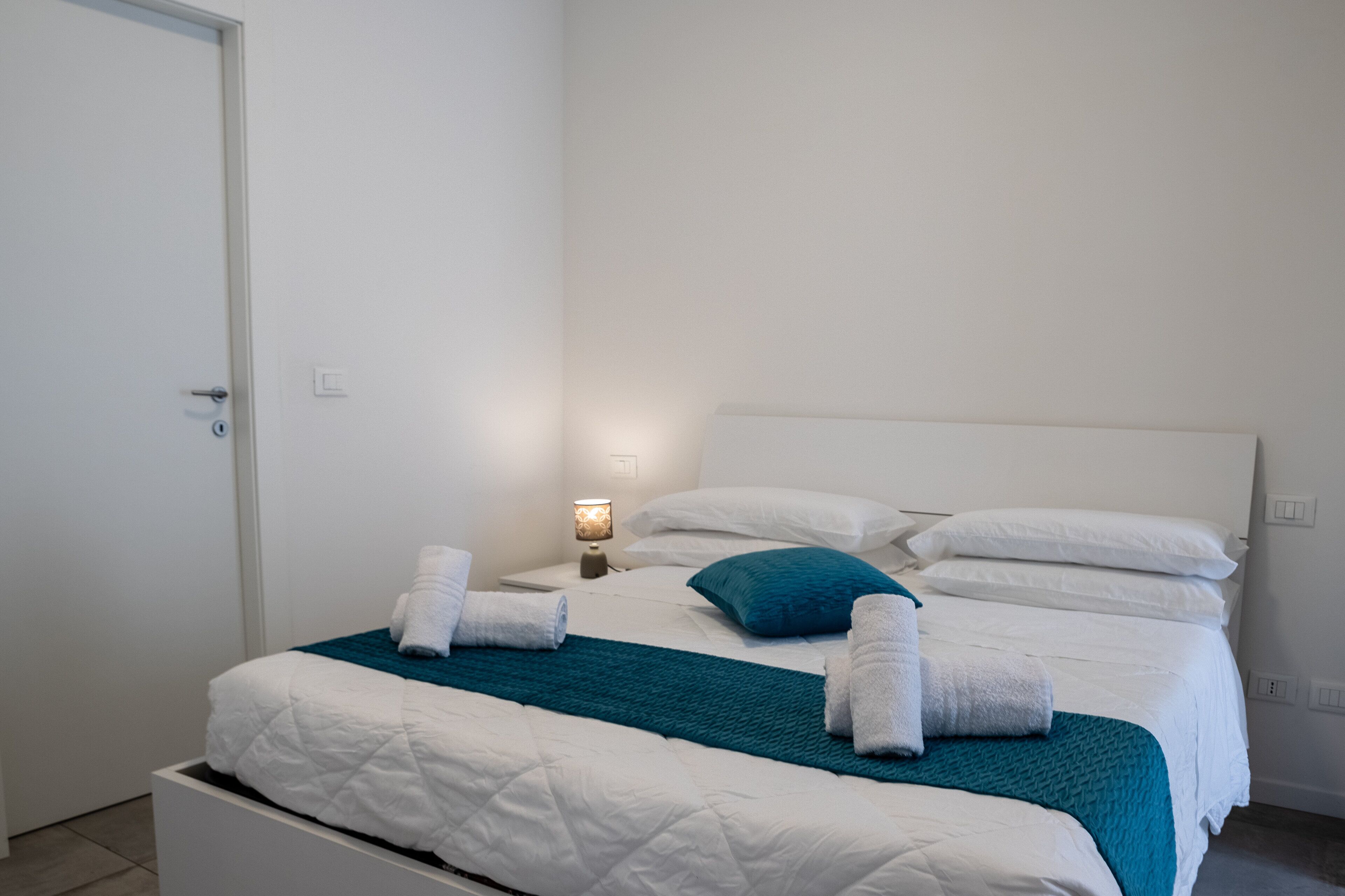 A neatly made bed with white and teal bedding, a bedside lamp, and towels on the bed.