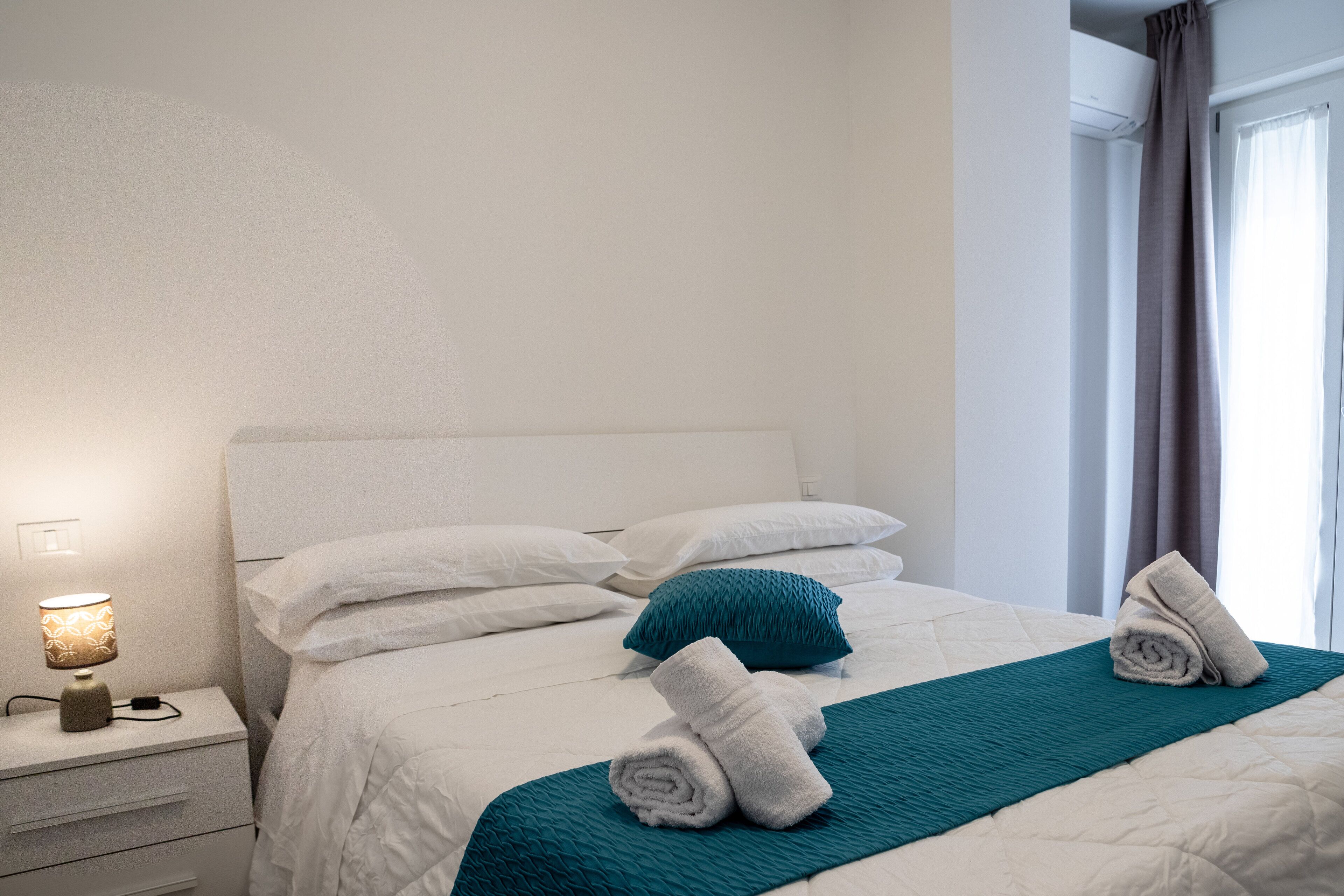 A neatly made bed with white bedding, a teal throw blanket, and rolled towels.