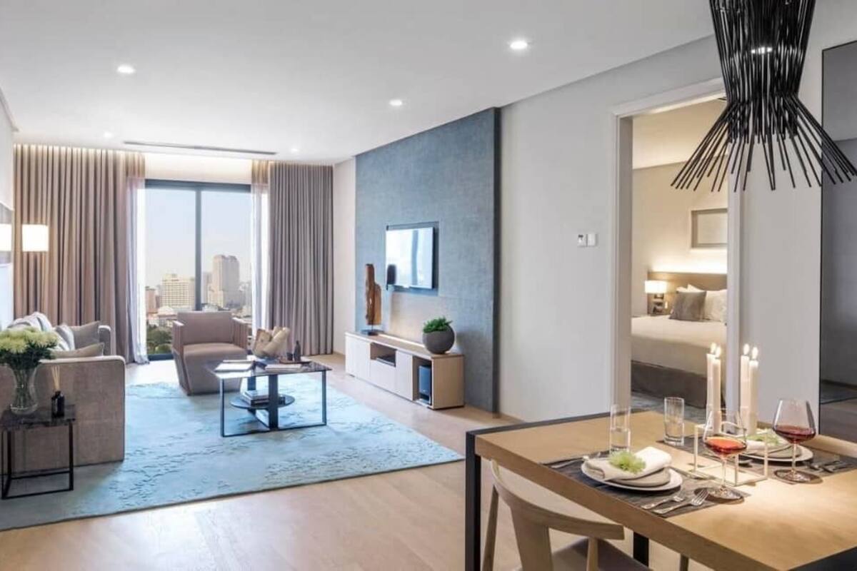 A modern hotel room with a dining area, a sofa set, a TV mounted on the wall, and a city view.