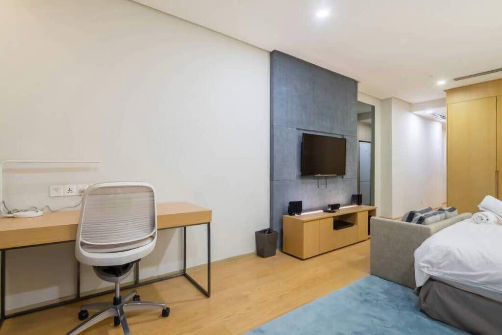 Ein modernes Hotelzimmer mit Schreibtisch aus Holz, einem Sessel, einem Flachbildfernseher an einer grauen Wand, einem Bett mit grauem Kopfteil und einer kleinen Sitzecke mit Kissen.