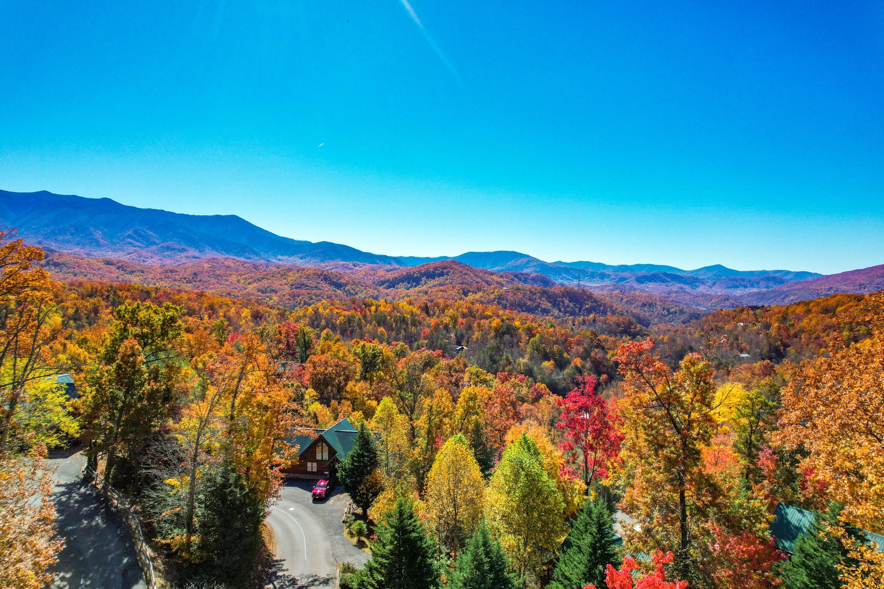 A cabin surrounded by a forest with autumn foliage, a winding road, and distant mountains.