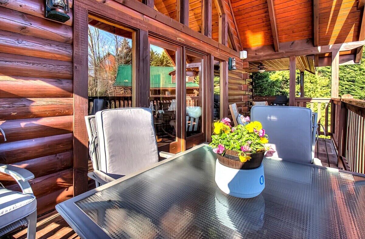 A wooden deck with a glass table and chairs, a potted plant, and a view of a cabin in the background.