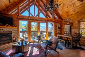 A spacious cabin interior with a fireplace, comfortable seating, and a large window offering a mountain view. - Cabin On A Cloud (Gatlinburg)