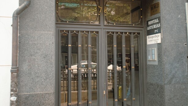 Entrada de hotel con una puerta metálica, cartel que indica tarifas de habitaciones y un coche estacionado afuera.