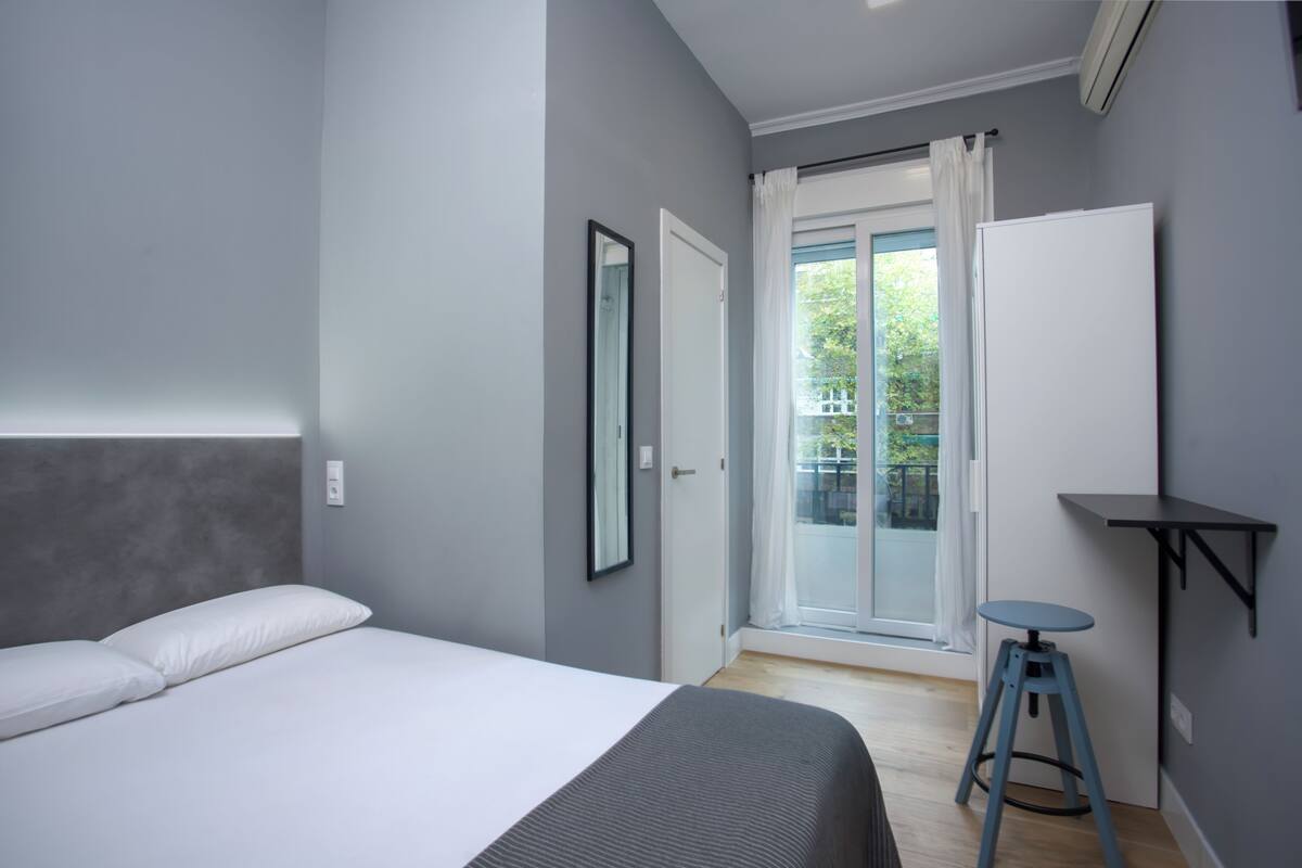 Un dormitorio moderno con cama, un taburete pequeño, una pizarra y un ventanal corredero a un balcón.