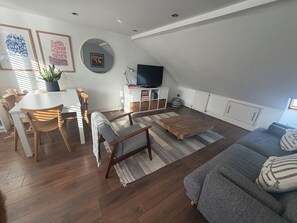 A modern living room with a dining area, wooden flooring, a grey sofa, and a TV on a white cabinet. - Cozy and Convenient Raynes Park maisonette (Greater London)