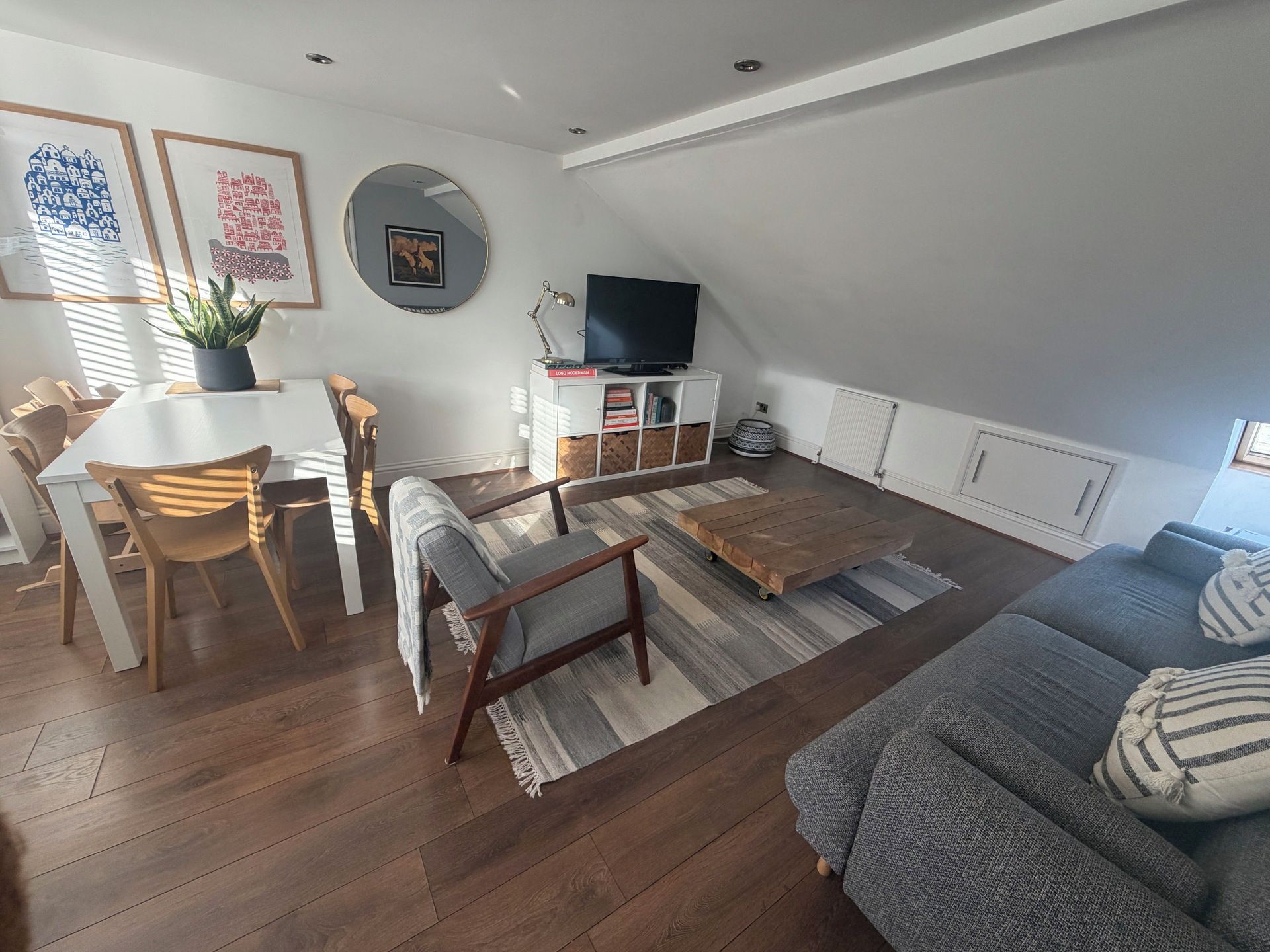 Un salon moderne comprenant un coin repas, un parquet, un canapé gris et un téléviseur posé sur un meuble blanc.