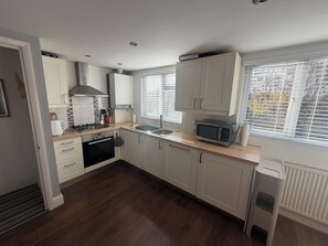 A modern kitchen with white cabinets, a gas stove, an oven, a microwave, and a window with blinds. - Cozy and Convenient Raynes Park maisonette (Greater London)