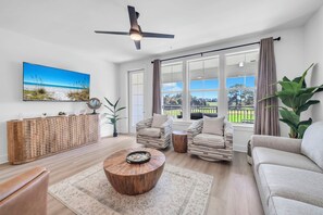 Ein geräumiges Wohnzimmer mit großem Fenster, Deckenventilator, Holztisch und einer Couch.