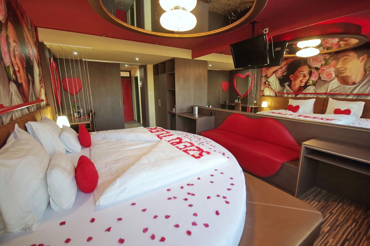 A hotel room with a bed, red chairs, a TV, and a wall with heart-shaped decorations.