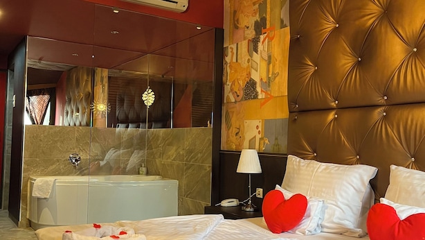 A hotel room with a large bed, a tufted headboard, a modern chandelier, a bathroom with a bathtub, and a mirror with a decorative frame.