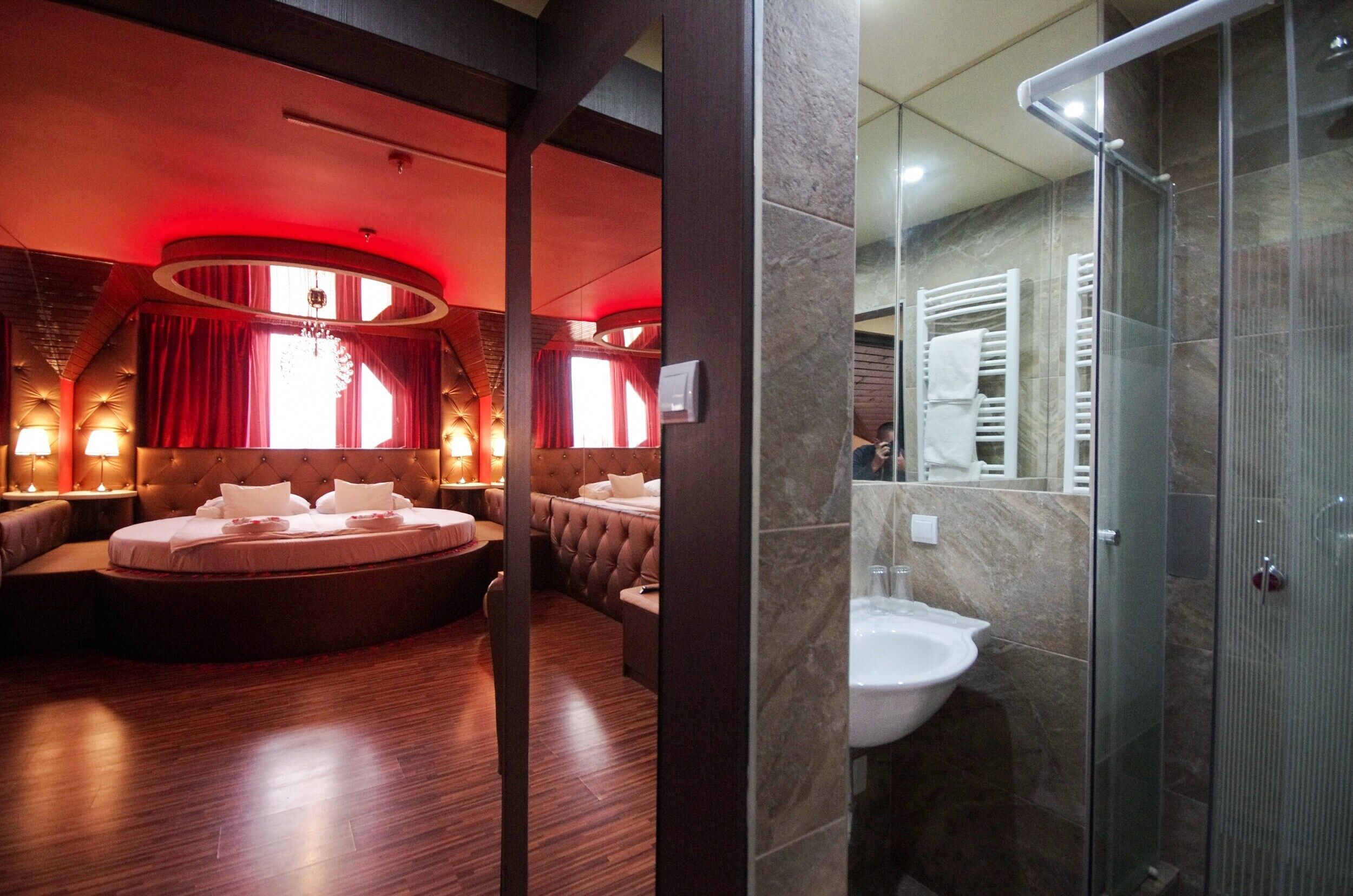 A hotel room with a circular seating area, a large bed, a separate bathroom with a glass shower, and a red-lit ambiance.
