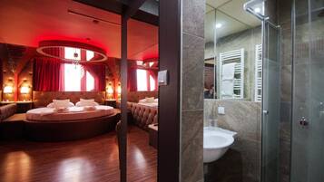 A hotel room with a circular seating area, a large bed, a separate bathroom with a glass shower, and a red-lit ambiance.