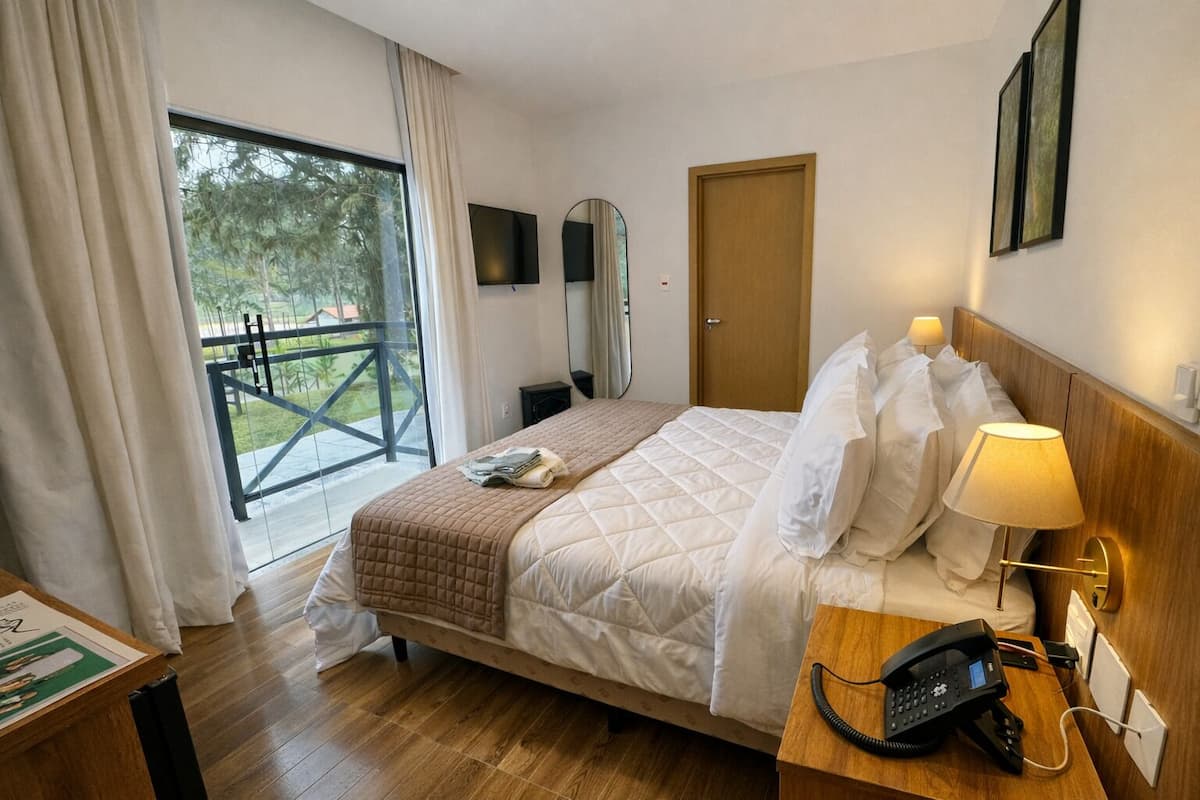 Ein Hotelzimmer mit Bett, Nachttischlampe, Telefon und Blick ins Freie durch ein großes Fenster.