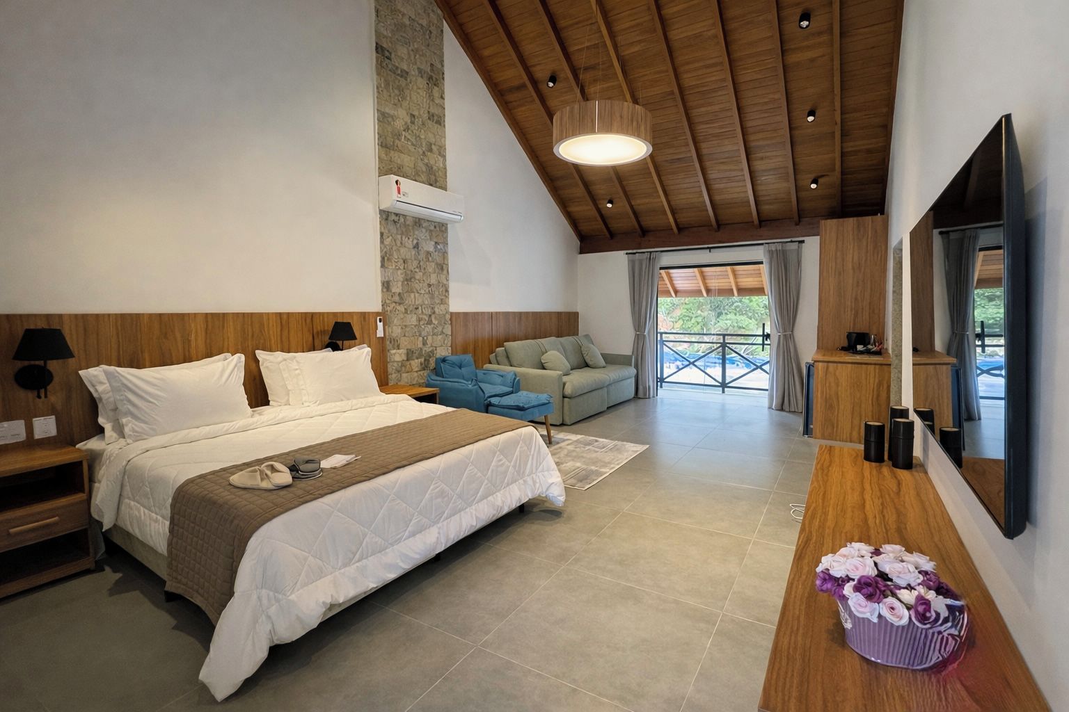 Un dormitorio moderno con una cama grande, techo de madera, pared de piedra, un sofá y un televisor.