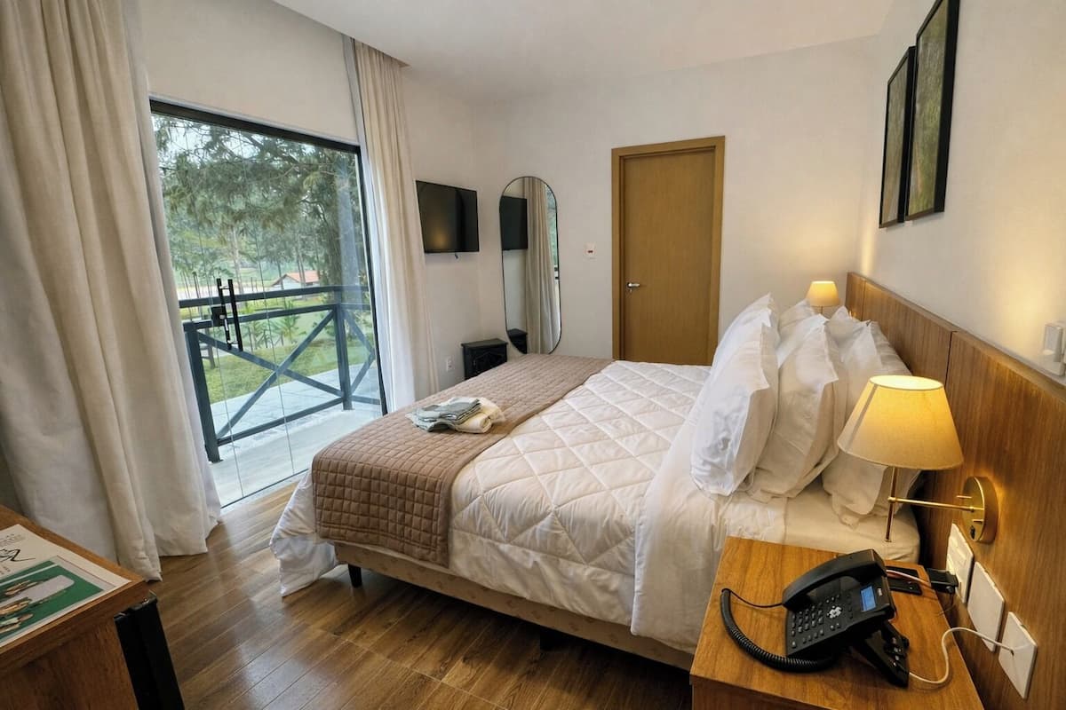 Ein Hotelzimmer mit einem großen Bett, einem Nachttisch mit Lampe, einem Telefon und Blick auf einen Balkon mit Geländer.