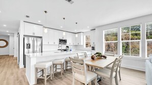 A modern kitchen with white cabinetry, a central island, and a dining area with light-colored chairs. - The Grove Dogwood 321 - 4 Bedrooms - Lazy River - Seascape Resort (Miramar Beach)