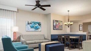Ein modernes Wohnzimmer mit grauem Sofa, Türkis-Armstühlen, einem blauen Hocker, einem Esstisch mit Stühlen und einem Deckenventilator.