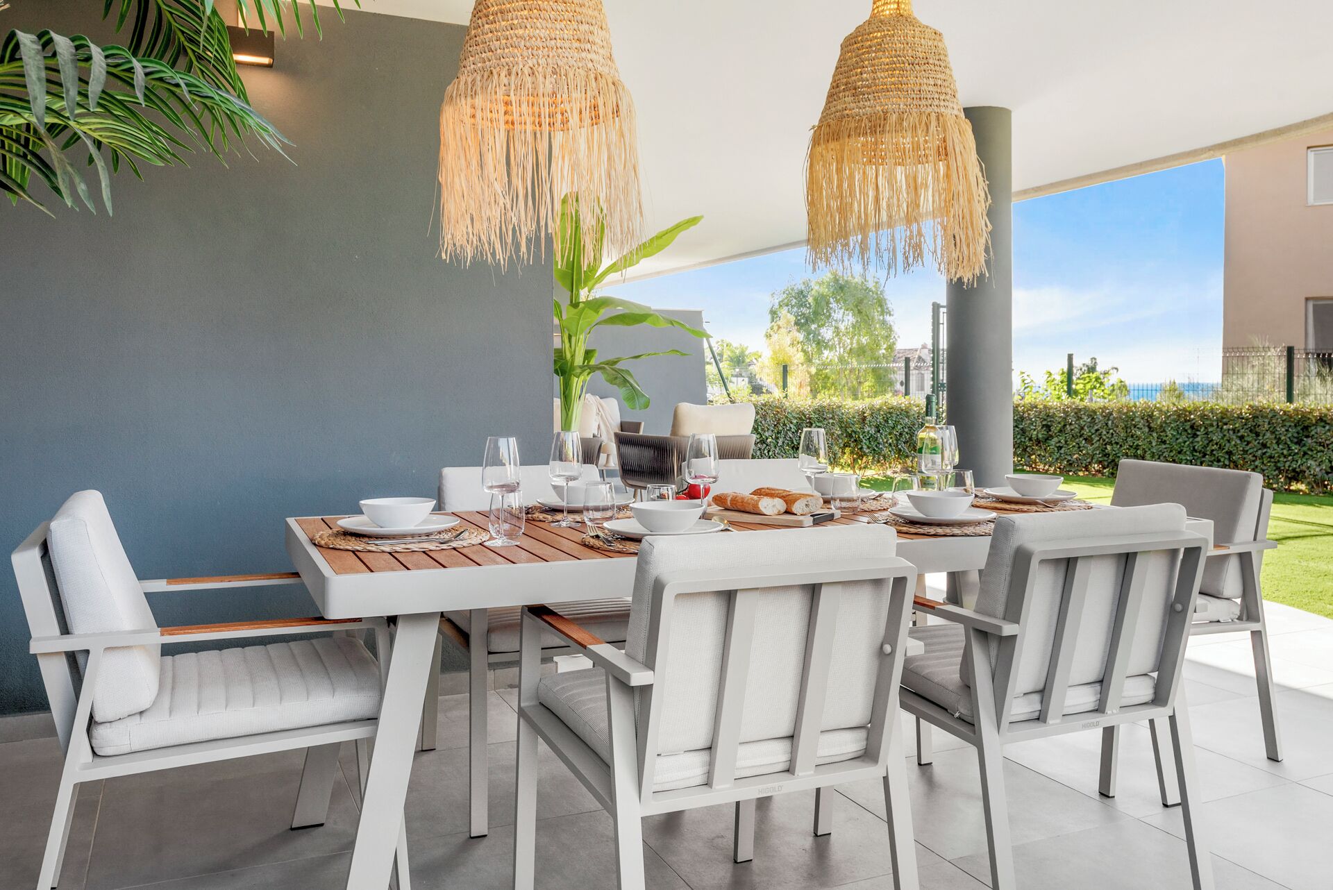 A modern outdoor dining area with a wooden table, white chairs, and woven pendant lights.