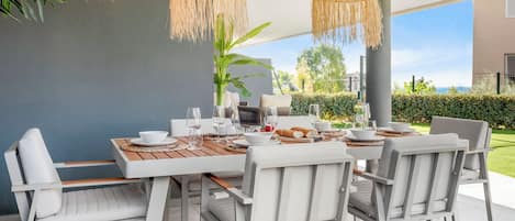 A modern outdoor dining area with a wooden table, white chairs, and woven pendant lights.