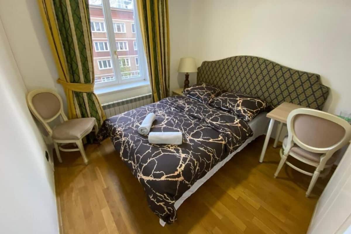 A small, neatly arranged bedroom with a bed, two chairs, a table, and a window with curtains.