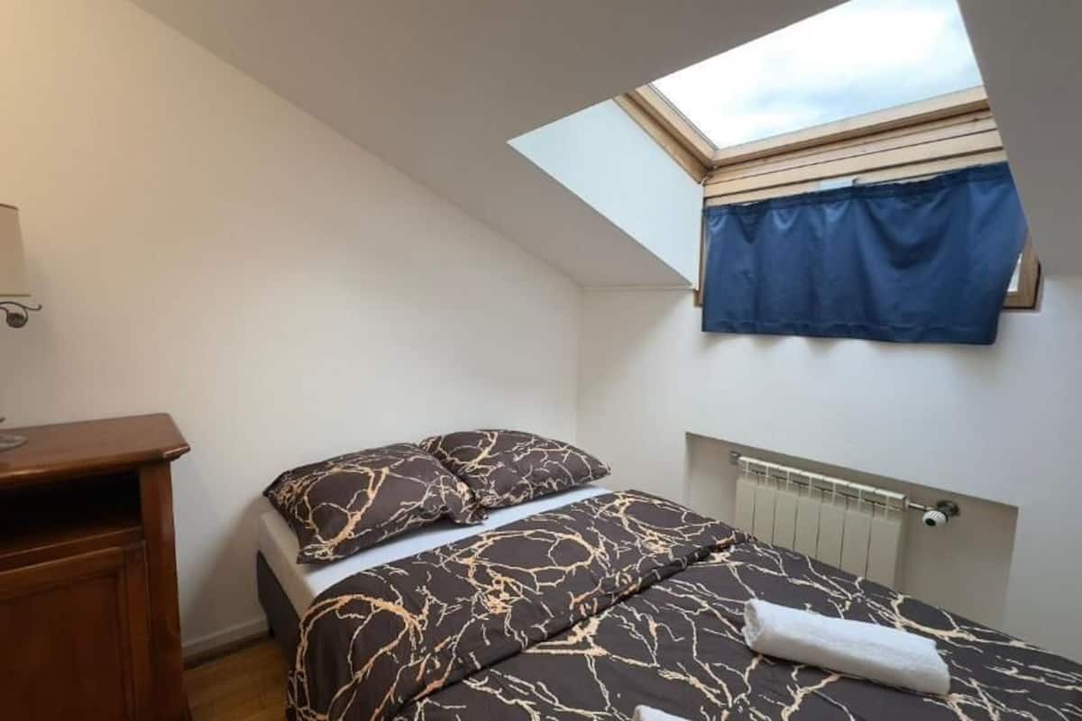 A bedroom with a skylight, a bed with patterned bedding, a bedside table with a lamp, and a radiator.