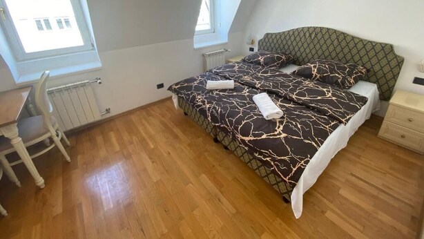 Ein Schlafzimmer mit schrägverdeckter Decke, einem Bett mit gemusterten Textilien, Holzboden, Schreibtisch mit Stuhl und einem Fenster mit Blick auf Gebäude.