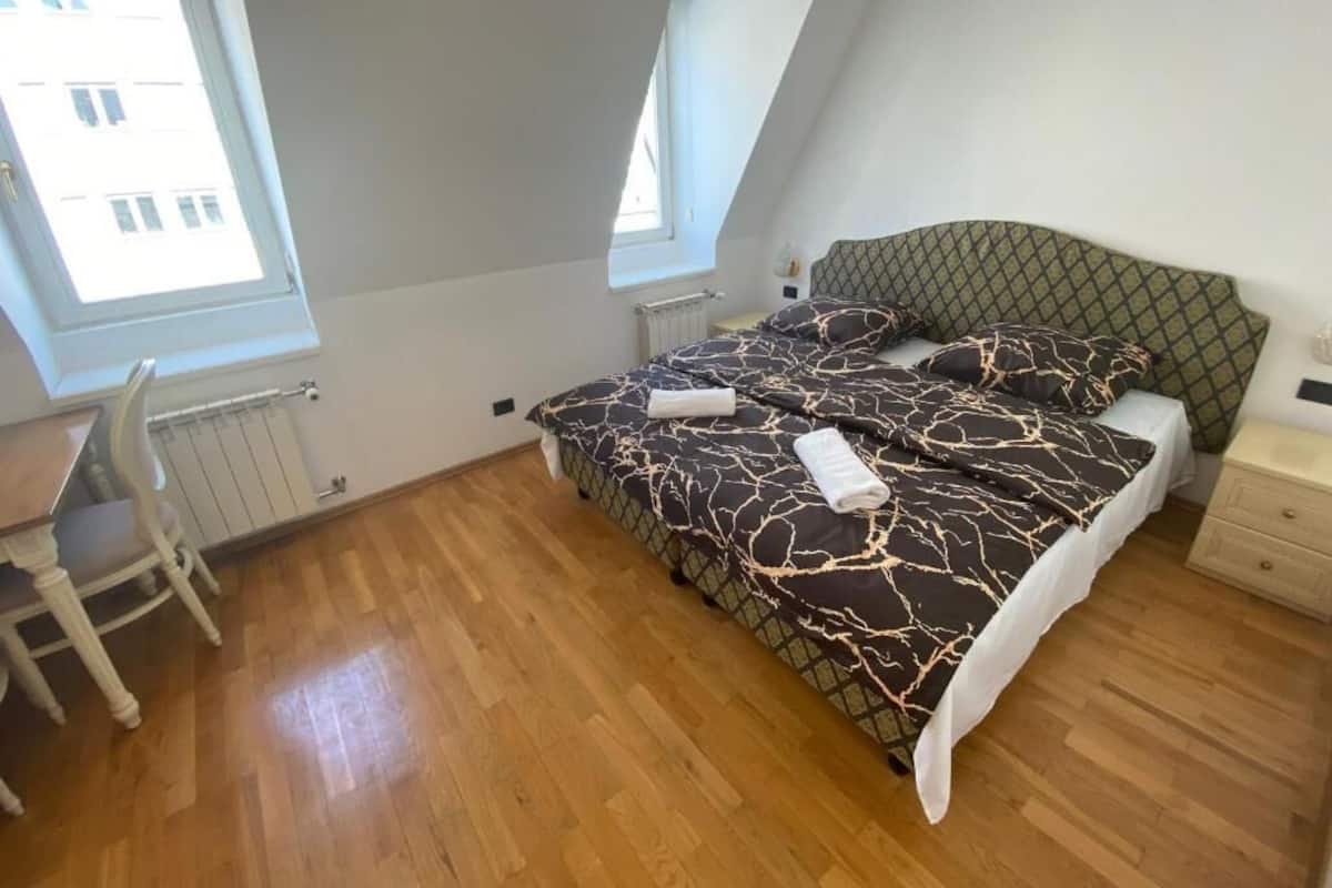 A bedroom with a sloped ceiling, a bed with patterned bedding, a wooden floor, a desk with a chair, and a window with a view of buildings.