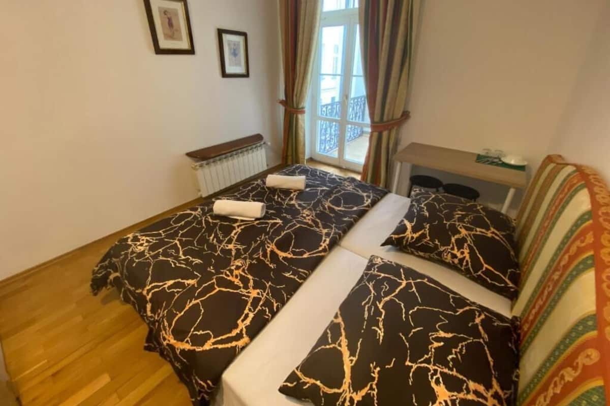 A bedroom with a bed, a radiator, a window with curtains, and a small table with a lamp.