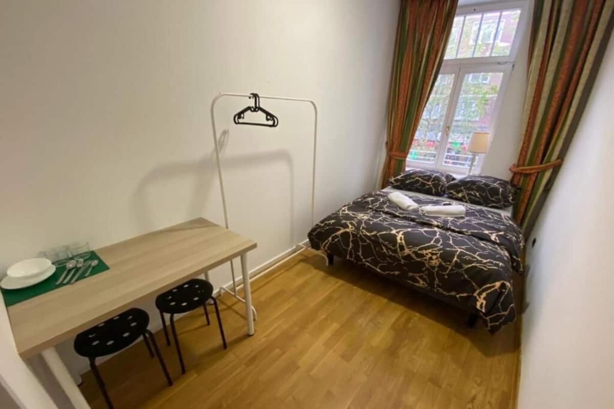 A small, tidy room with a bed, a table with chairs, and a window with curtains.
