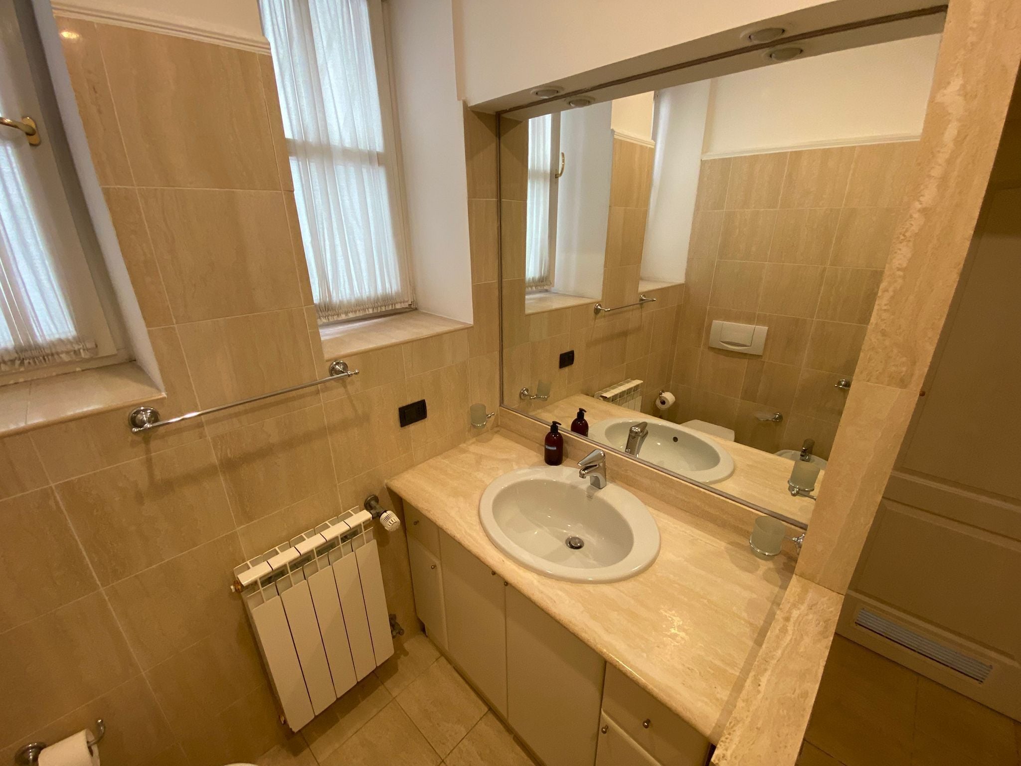 A modern bathroom with two sinks, a large mirror, a window with curtains, and a radiator.