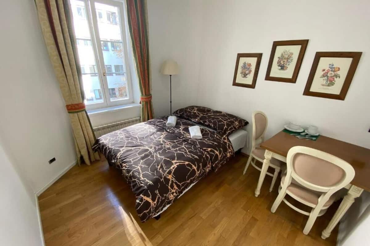 A small, tidy room with a bed, a desk, and a chair. There are three framed pictures on the wall.