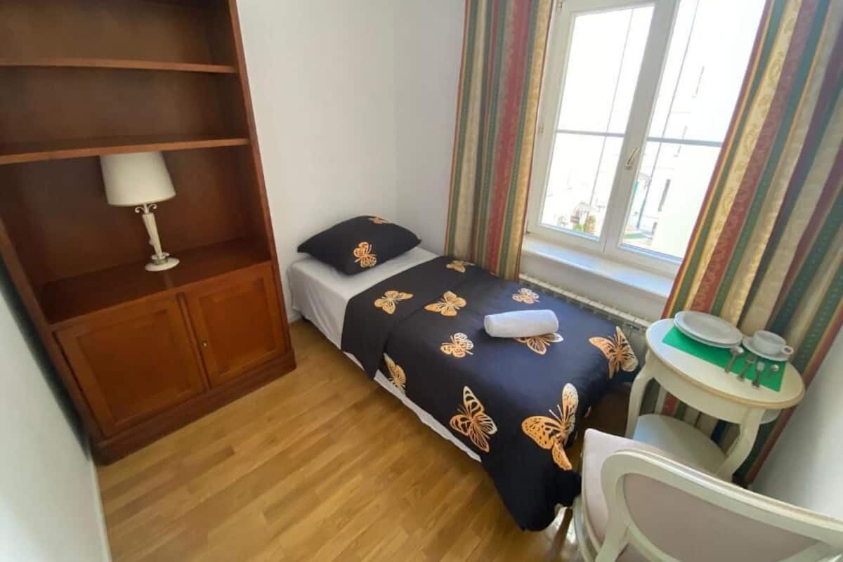 A small bedroom with a bed, a bedside table, a chair, a wardrobe, and a window with curtains.
