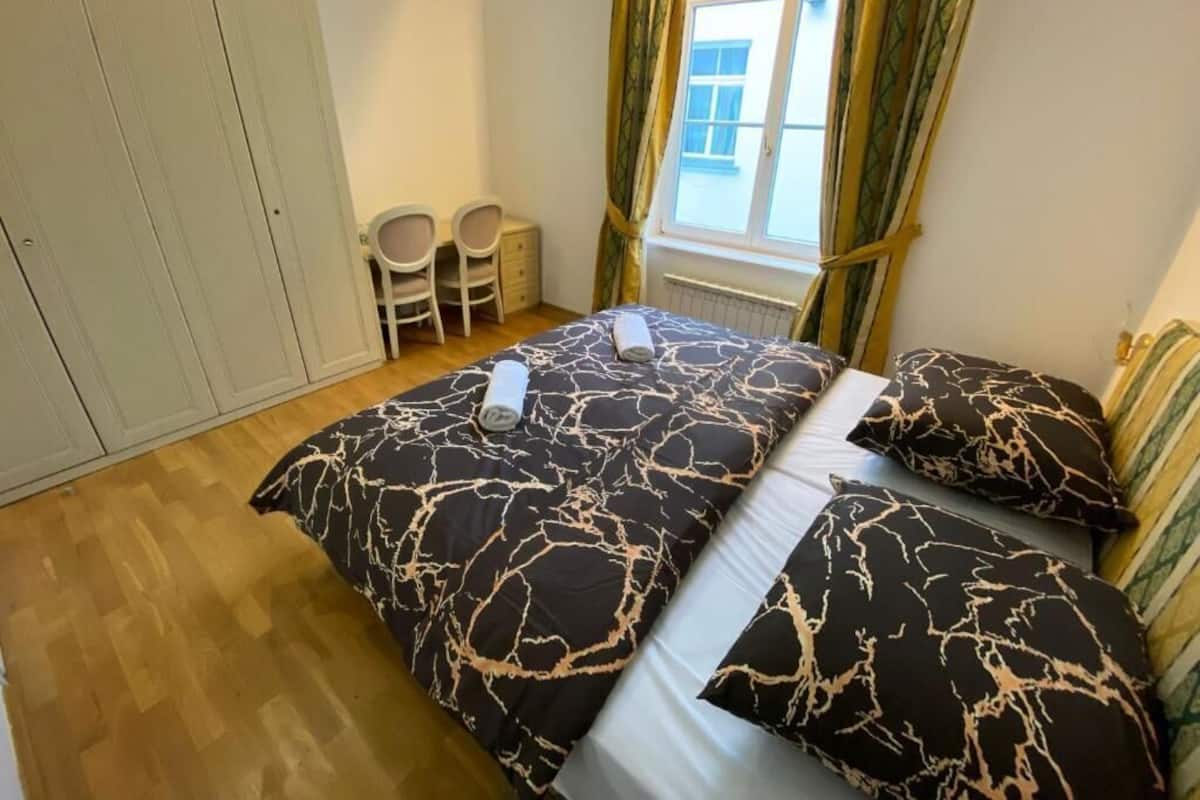 A bedroom with a bed, two chairs, a window with curtains, and a radiator.