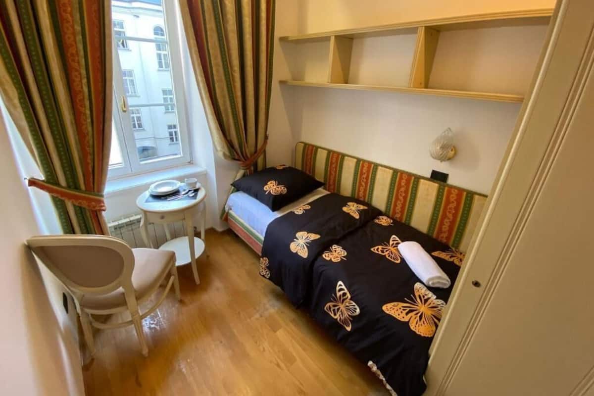 A small, compact room with a single bed, a chair, a small table, and a window with curtains.