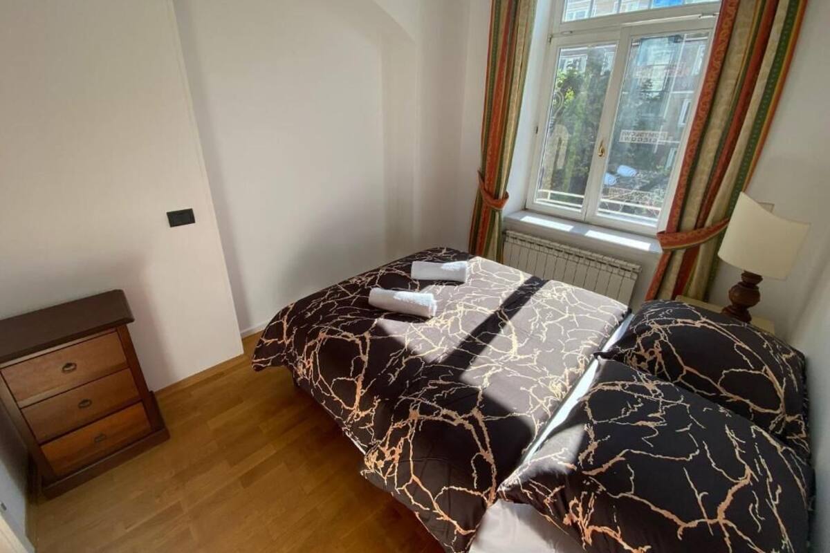 A bedroom with a bed, a nightstand, a window with curtains, and a radiator.