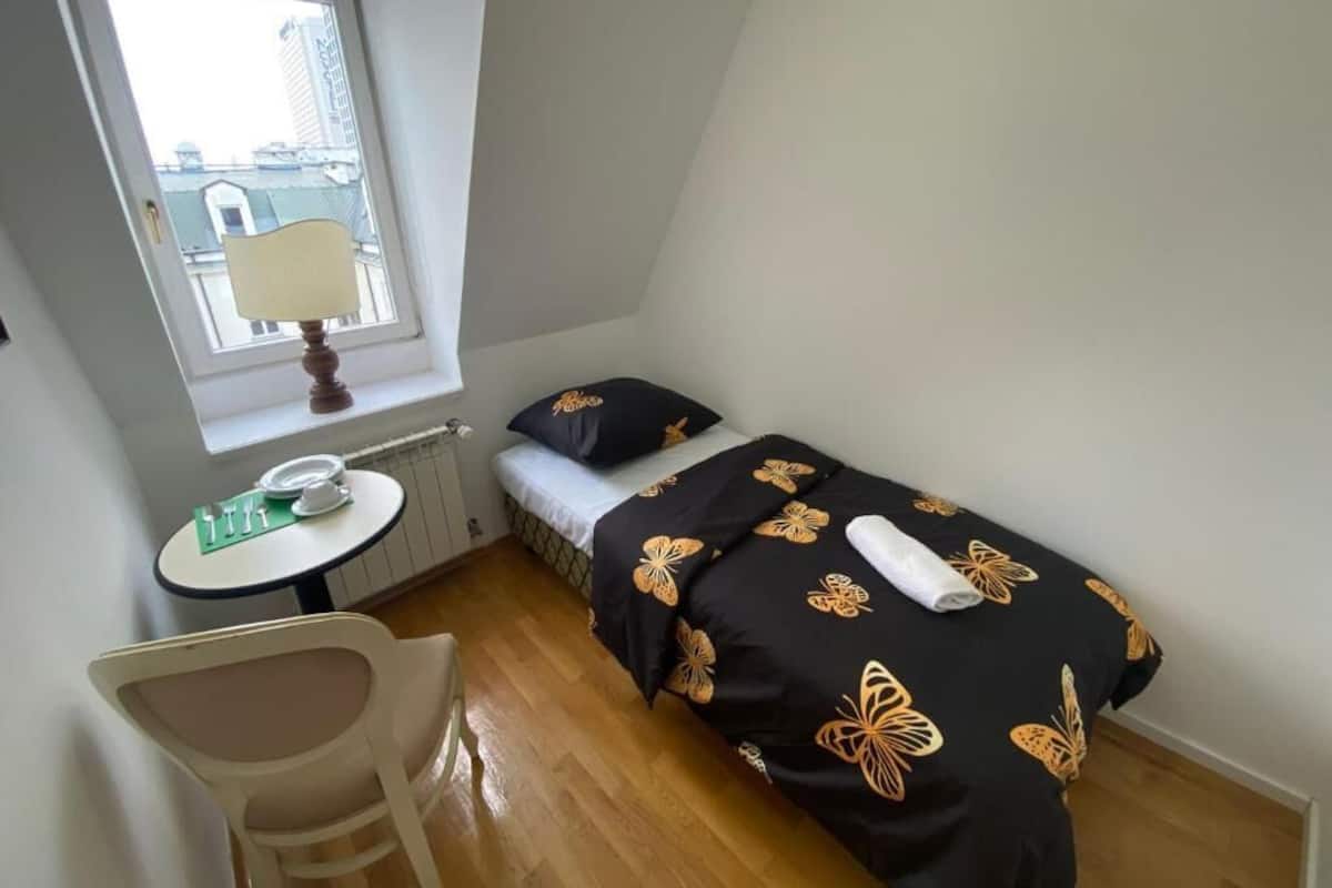 A small, single-room apartment with a bed, a small table, and a chair. There is a window providing natural light and a view of buildings outside.
