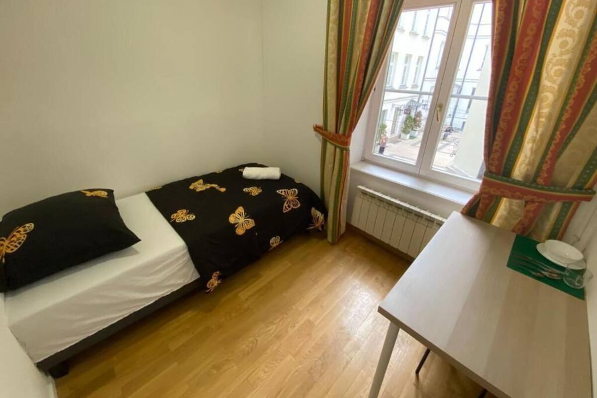 A small, single-bed room with a wooden floor, a window with curtains, and a table with a plate and cup.
