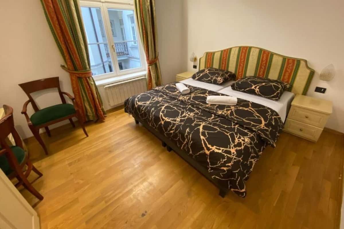 A bedroom with a bed, wooden floor, a chair, a nightstand, and a window with curtains.