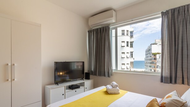 A hotel room with a bed, a TV on a white cabinet, a window with curtains, and a view of buildings and the sea. - 300 Meters From Copacabana Beach Nsc909 (Rio de Janeiro)