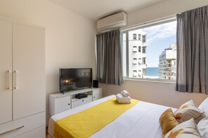 Una habitación de hotel con una cama, un televisor sobre un mueble blanco, una ventana con cortinas y vistas a edificios y al mar.