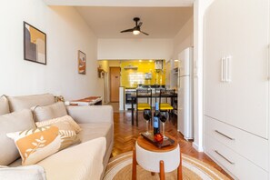 Una sala de estar moderna con un sofá, una mesa de centro con copas de vino y una cocina al fondo con sillas amarillas.
