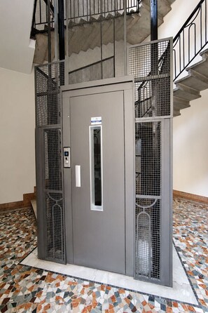 Un ascensore grigio con un display digitale, circondato da un pavimento piastrellato e ringhiere in metallo.