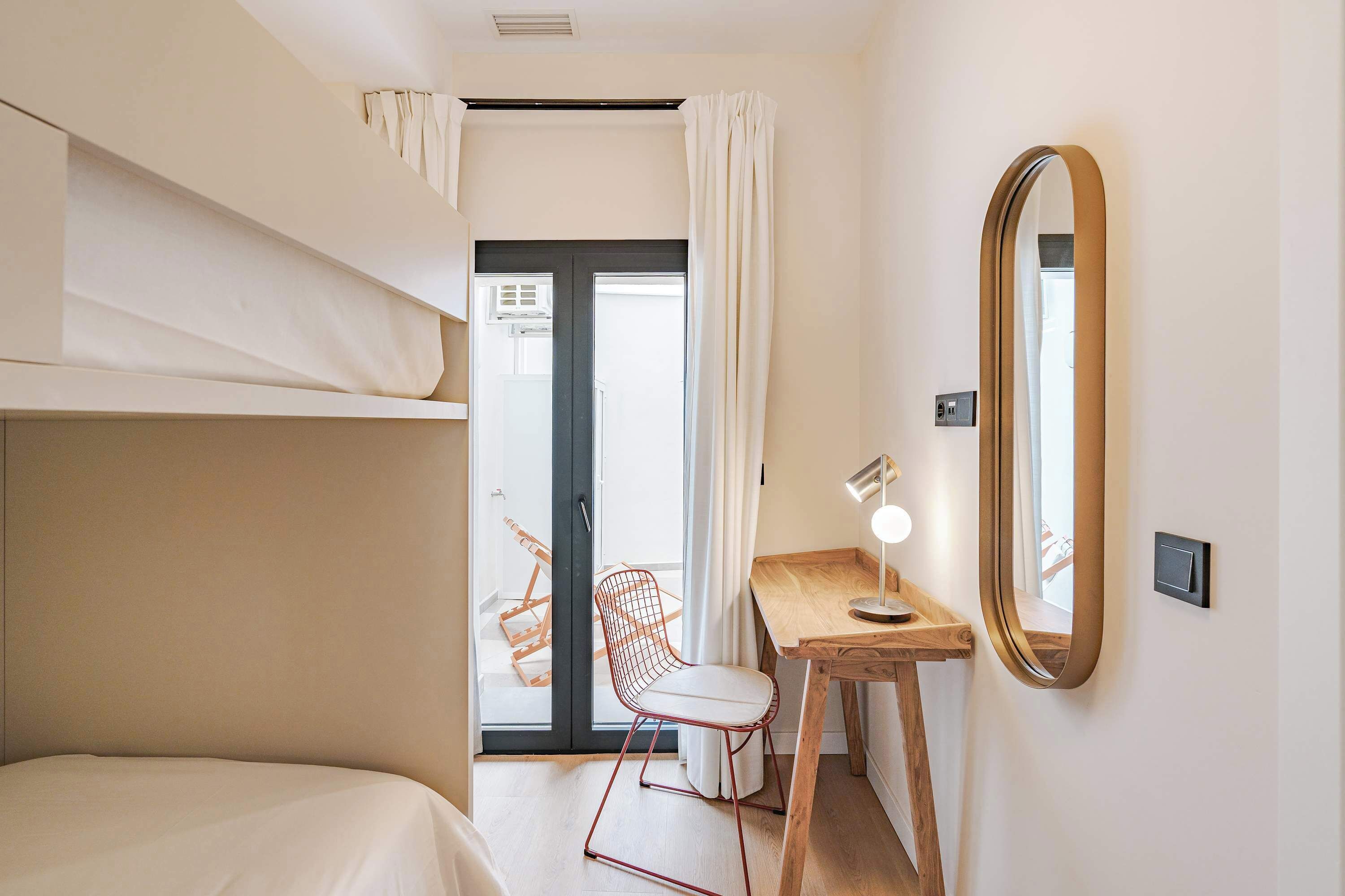 Una habitación pequeña y bien iluminada con una cama litera, un escritorio con silla, un espejo y un puertas corredera de vidrio que da a un espacio exterior.