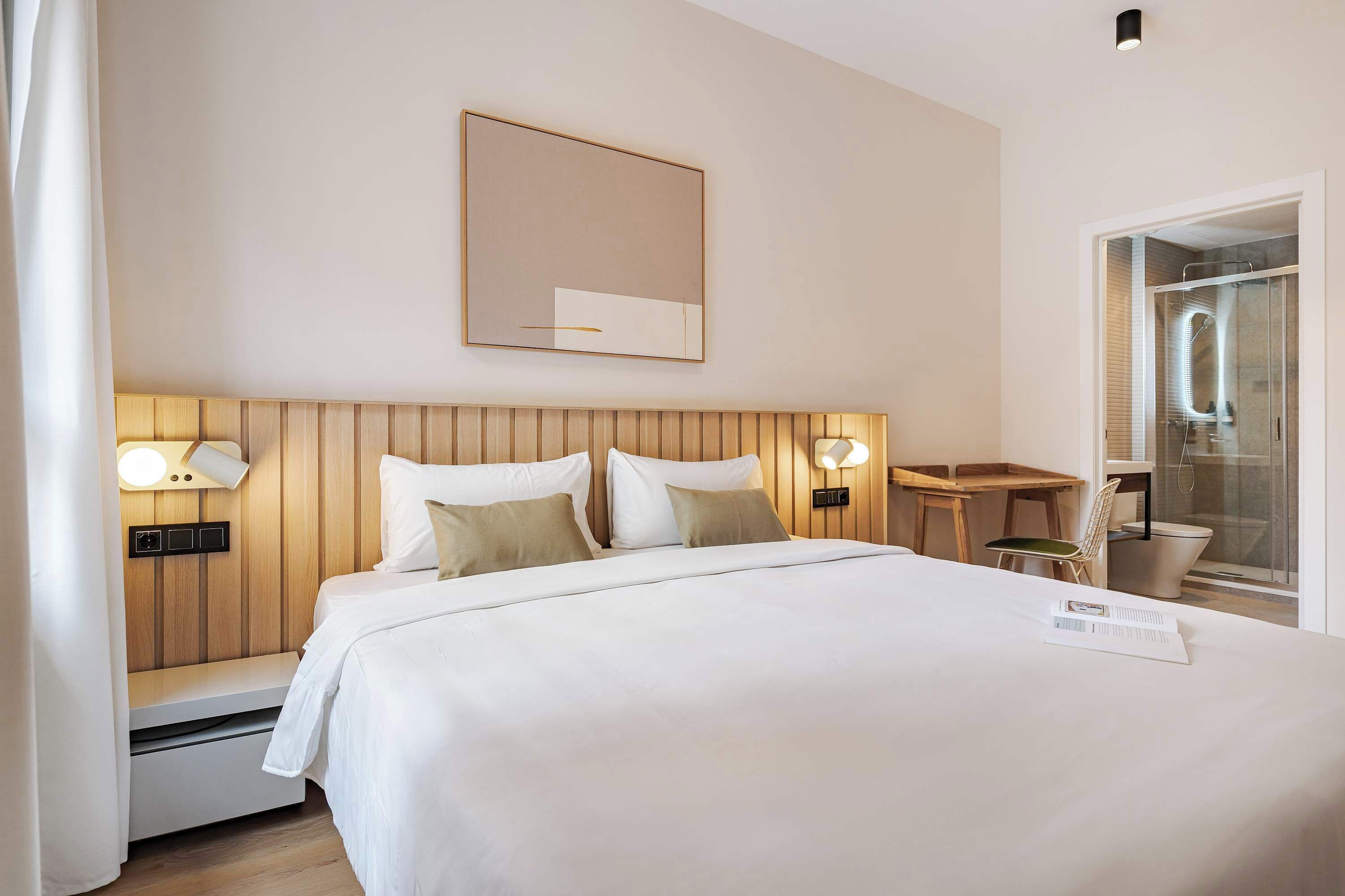 A hotel room with a bed, bedside lamps, a mirror, a small table, and a shower area.