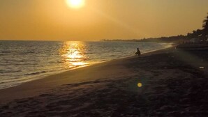 Una playa al atardecer, con el sol bajo en el horizonte, mar tranquilo y algunas personas caminando por la orilla.