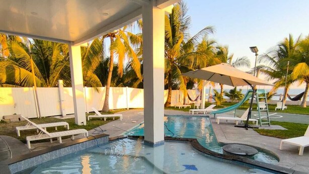 Una piscina con sillas de descanso y un tobogán, rodeada de palmeras y una valla blanca.